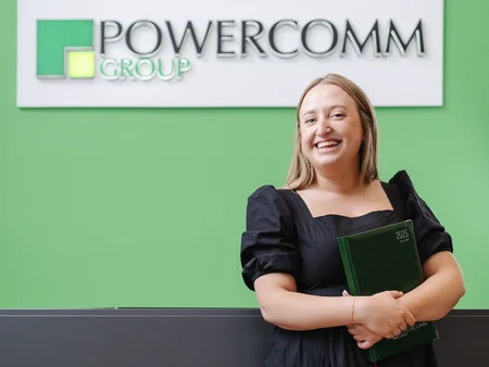 A smiling blonde woman standing in front of reception desk that has a large sign that says Powercomm Group