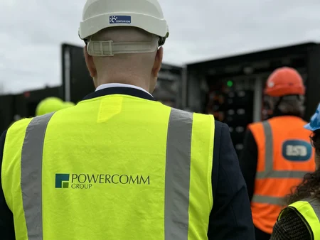 Martin, with his back to the camera, wears a high-vis jacket with the Powercomm logo looking at a Battery Storage solution unit. 