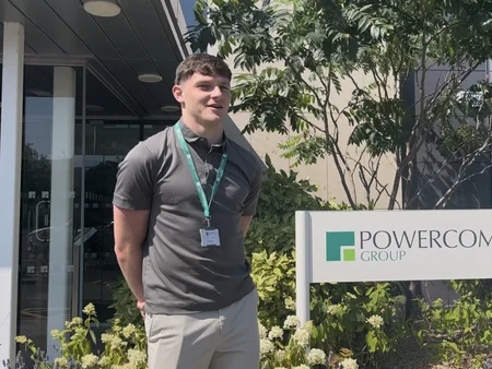 A young man standing outside in the sun, smiling beside the Powercomm Group sign