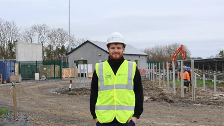 Young man with hardhat and high viz, standing in front of busy construction site