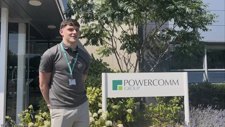 A young man standing outside in the sun, smiling beside the Powercomm Group sign